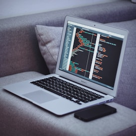 A laptop sits on a gray fabric couch, displaying code on its screen. Next to it, a smartphone lies flat. The code appears to be written in HTML or a similar programming language.