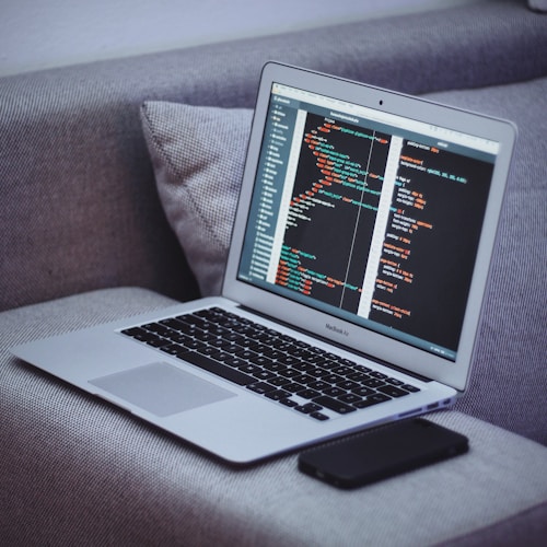 A laptop sits on a gray fabric couch, displaying code on its screen. Next to it, a smartphone lies flat. The code appears to be written in HTML or a similar programming language.