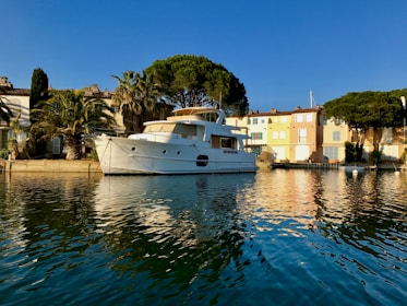 A large white yacht is moored alongside a quai in a serene coastal town. The scene is framed by palm trees and colorful buildings in soft pastel shades, bathed in warm sunlight. The water reflects the vivid colors of the buildings and the deep blue of the sky.