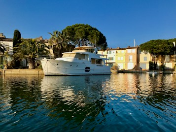 A large white yacht is moored alongside a quai in a serene coastal town. The scene is framed by palm trees and colorful buildings in soft pastel shades, bathed in warm sunlight. The water reflects the vivid colors of the buildings and the deep blue of the sky.