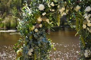An archway beautifully adorned with lush greenery and a variety of white and pale-colored flowers, with a calm, reflective body of water in the background under a sunlit sky, creating a picturesque and serene scene.