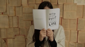 A person is holding an open book in front of their face, obscuring their identity. The book displays a message that encourages experiencing life instead of merely existing. The background is composed of open books, creating an artistic or literary setting. The person is wearing a light-colored sweater.