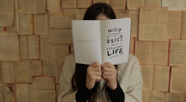 A person is holding an open book in front of their face, obscuring their identity. The book displays a message that encourages experiencing life instead of merely existing. The background is composed of open books, creating an artistic or literary setting. The person is wearing a light-colored sweater.