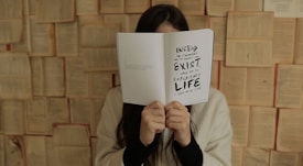 A person is holding an open book in front of their face, obscuring their identity. The book displays a message that encourages experiencing life instead of merely existing. The background is composed of open books, creating an artistic or literary setting. The person is wearing a light-colored sweater.