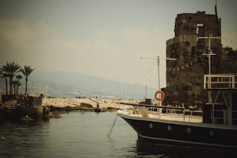 A tranquil harbor scene featuring a classic wooden boat with a dark hull floating on calm water. To the right, a rustic, ancient stone tower stands with hanging string lights. In the background, palm trees and a rocky pier can be seen, along with a distant hilly landscape shrouded in haze.