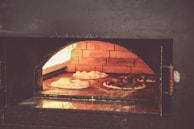 A brick pizza oven with several pizzas baking inside. The interior of the oven glows warmly from the fire, highlighting the rustic texture of the bricks. The pizzas are placed on the oven's stone surface, with toppings beginning to cook and dough rising.