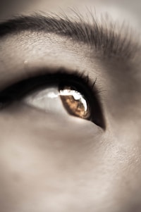 A close-up of a human eye with visible eyelashes and a reflection in the pupil. The eyebrow is partially visible at the top of the image. The lighting creates a soft, detailed texture on the skin around the eye.
