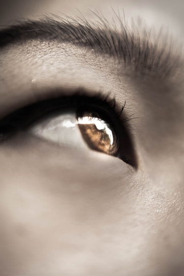 A close-up of a human eye with visible eyelashes and a reflection in the pupil. The eyebrow is partially visible at the top of the image. The lighting creates a soft, detailed texture on the skin around the eye.