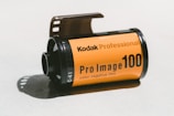 A roll of Kodak Professional Pro Image 100 color negative film on a light surface. The film canister is predominantly orange with black text and details. The film is partially extended from the canister.