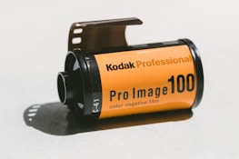 A roll of Kodak Professional Pro Image 100 color negative film on a light surface. The film canister is predominantly orange with black text and details. The film is partially extended from the canister.