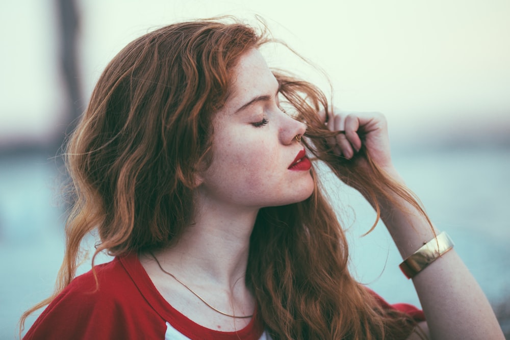 A woman with long red hair and red lipstick holds her hair while closing her eyes