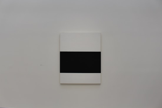 A minimalist art piece hangs on a plain light gray wall. The artwork features a large black rectangle centered on a white canvas.