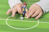 A child's hands are holding a toy figurine dressed in a blue and yellow sports uniform on a miniature soccer field. A small soccer ball is positioned next to the toy, centered in a white circle marking on the green field.