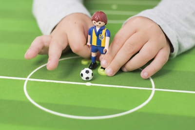 A child's hands are holding a toy figurine dressed in a blue and yellow sports uniform on a miniature soccer field. A small soccer ball is positioned next to the toy, centered in a white circle marking on the green field.