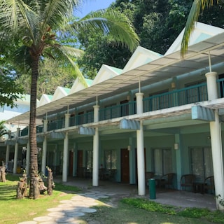 A two-story building with a row of rooms, featuring large windows and a covered balcony. Palm trees and lush greenery surround the building, and there is a sunny, tropical ambiance.