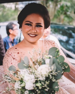 A woman with a warm smile is holding a bouquet of flowers, adorned in a shimmering outfit with sequins and crystals. The background includes blurred figures and greenery, creating a lively and cheerful atmosphere.
