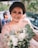 A woman with a warm smile is holding a bouquet of flowers, adorned in a shimmering outfit with sequins and crystals. The background includes blurred figures and greenery, creating a lively and cheerful atmosphere.