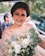 A woman with a warm smile is holding a bouquet of flowers, adorned in a shimmering outfit with sequins and crystals. The background includes blurred figures and greenery, creating a lively and cheerful atmosphere.