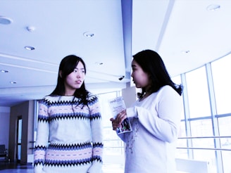 Two women are standing in a modern, well-lit space with large windows. One woman, wearing a patterned sweater, is looking towards the other woman, who holds a brochure in her hands. The atmosphere seems casual and conversational.