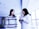 Two women are standing in a modern, well-lit space with large windows. One woman, wearing a patterned sweater, is looking towards the other woman, who holds a brochure in her hands. The atmosphere seems casual and conversational.