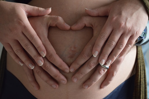 Hands forming a heart shape over a pregnant belly, with one set of hands wearing a ring and a watch visible on the wrist. The hands are gently placed, indicating a tender and caring gesture.