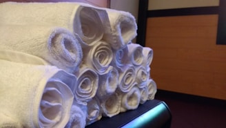 A neatly arranged stack of rolled white towels sitting on a surface. The background features a wooden panel wall with some dark edges.