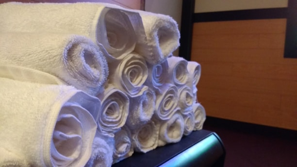 A neatly arranged stack of rolled white towels sitting on a surface. The background features a wooden panel wall with some dark edges.