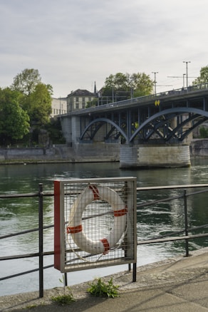 A riverbank scene features a metal railing with a lifebuoy encased in a wire mesh stand in the foreground. There's a bridge crossing the river, which reflects a mix of blue and gray tones on the water. In the background, leafy trees and historic-looking buildings add a scenic and serene touch to the environment.