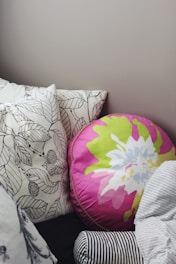A collection of pillows arranged on a bed or couch. One pillow is round with a vibrant floral design in pink, green, and white colors. Other pillows feature black and white leaf patterns, while another has black and white stripes. The setting appears cozy and inviting.