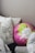A collection of pillows arranged on a bed or couch. One pillow is round with a vibrant floral design in pink, green, and white colors. Other pillows feature black and white leaf patterns, while another has black and white stripes. The setting appears cozy and inviting.