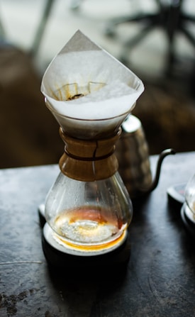 A Chemex coffee maker sits on a dark surface with freshly brewed coffee visible in the bottom. The paper filter, partly stained with coffee grounds, is positioned at the top. The surroundings are slightly blurred, focusing on the coffee maker.