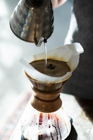 A hand pours hot water from a metal gooseneck kettle over a paper filter in a Chemex coffee maker. The coffee grounds in the filter are being saturated, with coffee dripping into the carafe below. The scene is lit with natural light, creating a warm and inviting atmosphere.