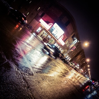 A nighttime urban scene with wet streets reflecting bright neon lights. A vintage theater marquee is prominently lit, and blurred cars pass by, creating a dynamic sense of motion. Streetlights illuminate the scene, and the overall atmosphere is reminiscent of a classic cityscape.