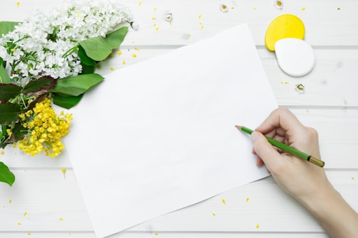 A hand holding a green colored pencil poised above a blank sheet of white paper, surrounded by white and yellow flowers on a light wooden surface, with scattered petals and small decorative objects.