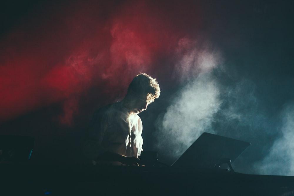 A musician in a white shirt on stage with white and red lights breaking through the smoke behind his back