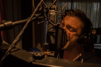 A person is singing passionately into a large microphone in a dimly lit recording studio. They are wearing headphones and are closely positioned to the microphone, emphasizing the intensity of their performance. The background features soundproofing curtains and studio equipment.