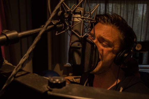 A person is singing passionately into a large microphone in a dimly lit recording studio. They are wearing headphones and are closely positioned to the microphone, emphasizing the intensity of their performance. The background features soundproofing curtains and studio equipment.