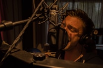 A person is singing passionately into a large microphone in a dimly lit recording studio. They are wearing headphones and are closely positioned to the microphone, emphasizing the intensity of their performance. The background features soundproofing curtains and studio equipment.