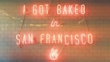 Neon sign with the text 'I GOT BAKED in SAN FRANCISCO' displayed on a tiled wall. The sign includes a peace hand symbol at the bottom.