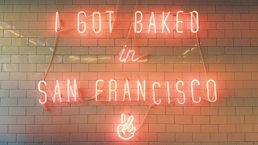 Neon sign with the text 'I GOT BAKED in SAN FRANCISCO' displayed on a tiled wall. The sign includes a peace hand symbol at the bottom.