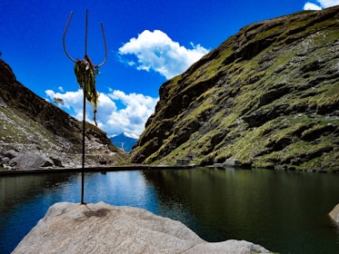 A picturesque mountainous landscape with a serene lake surrounded by steep, green hills. In the foreground, a trident is mounted on a rock, adorned with green and yellow drapes. The sky is vividly blue with scattered white clouds.