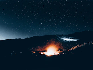 Campfire glowing warmly at night surrounded by tents nestled in a rugged mountain valley.