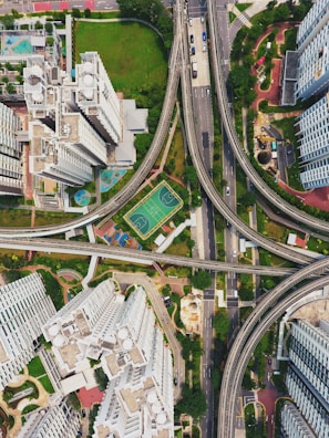 An aerial view of a modern urban landscape featuring high-rise buildings, intersecting roads, and elevated railways. In the center is a multi-use sports court surrounded by greenery. The architecture showcases a mix of residential and infrastructural elements, efficiently utilizing the available space.