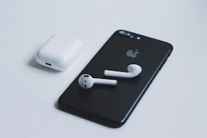 A black smartphone with a recognizable logo on the back is placed on a light surface. Next to it is a pair of white wireless earbuds, and their charging case is situated nearby. The composition highlights the sleek, minimalist design of the electronic devices.