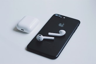 A black smartphone with a recognizable logo on the back is placed on a light surface. Next to it is a pair of white wireless earbuds, and their charging case is situated nearby. The composition highlights the sleek, minimalist design of the electronic devices.