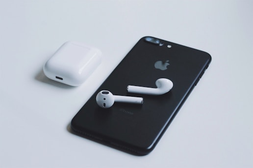 A black smartphone with a recognizable logo on the back is placed on a light surface. Next to it is a pair of white wireless earbuds, and their charging case is situated nearby. The composition highlights the sleek, minimalist design of the electronic devices.
