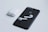 A black smartphone with a recognizable logo on the back is placed on a light surface. Next to it is a pair of white wireless earbuds, and their charging case is situated nearby. The composition highlights the sleek, minimalist design of the electronic devices.