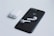 A black smartphone with a recognizable logo on the back is placed on a light surface. Next to it is a pair of white wireless earbuds, and their charging case is situated nearby. The composition highlights the sleek, minimalist design of the electronic devices.