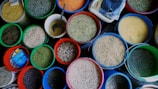 An assortment of various grains and seeds displayed in colorful plastic buckets. Each bucket is filled to the brim with different types of grains such as lentils, beans, and peas. A metal scoop is visible in one of the buckets, suggesting the grains are sold by weight. The vibrant colors of the buckets and the grains create an eye-catching display.