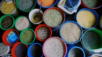 An assortment of various grains and seeds displayed in colorful plastic buckets. Each bucket is filled to the brim with different types of grains such as lentils, beans, and peas. A metal scoop is visible in one of the buckets, suggesting the grains are sold by weight. The vibrant colors of the buckets and the grains create an eye-catching display.